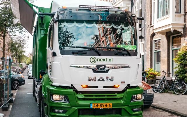 Een groen-witte vuilniswagen in een smalle straat met geparkeerde fietsen