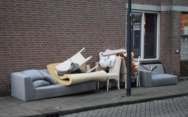 Een bank, een opgerold tapijt en stoelen aan de stoeprand bij een woning
